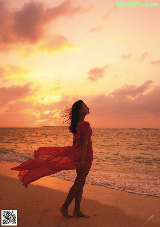 A woman in a red dress standing on the beach at sunset.